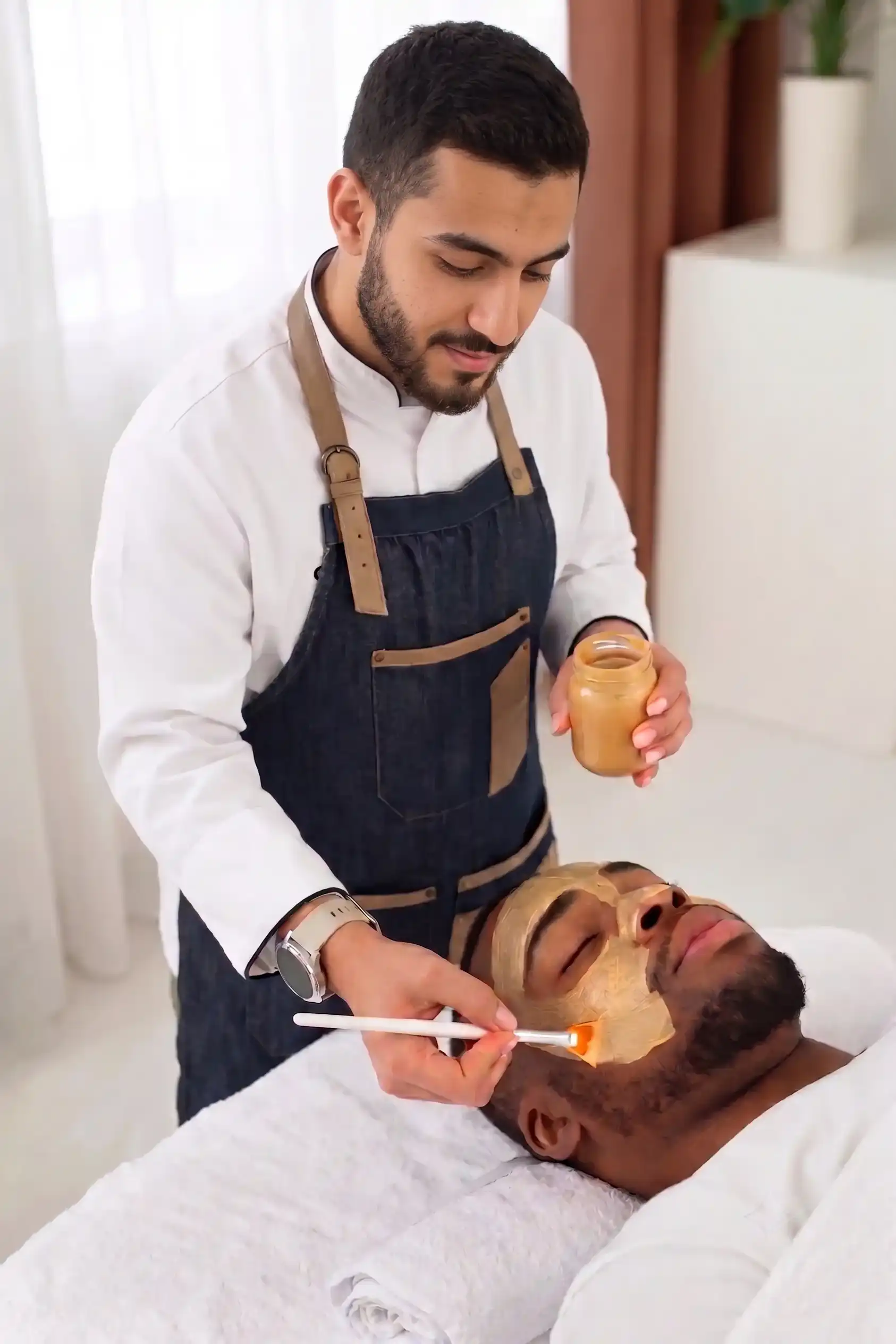 An aesthetician applying a golden facial mask to a male client’s face using a brush.