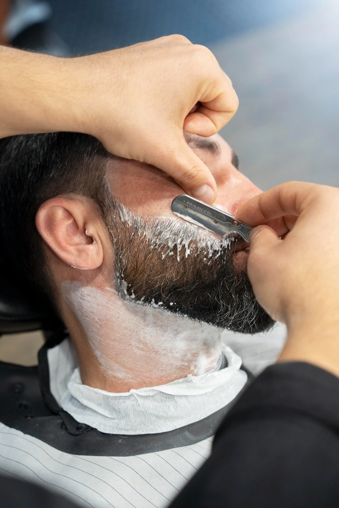 A professional barber applying shaving cream and using a straight razor for a beard grooming session.